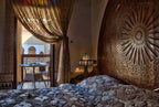 Riad Salam Fes bedroom with ornate carved wooden headboard, patterned blue comforter, decorative lamp, and balcony with seating