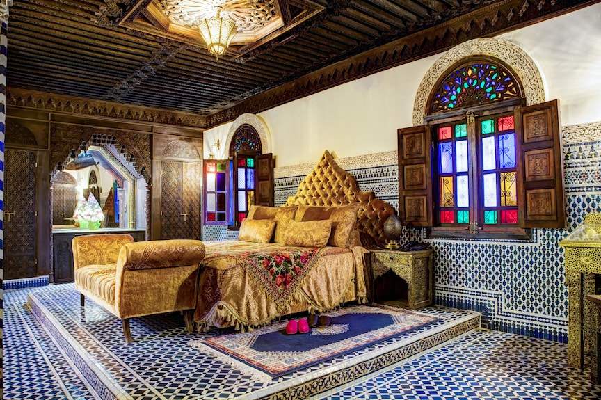 Riad Salam Fes bedroom with ornate wooden ceiling, colorful stained glass windows, patterned tile walls and floor, and gold velvet bedding