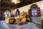 Riad Salam Fes bedroom with ornate wooden ceiling, colorful stained glass windows, patterned tile walls and floor, and gold velvet bedding