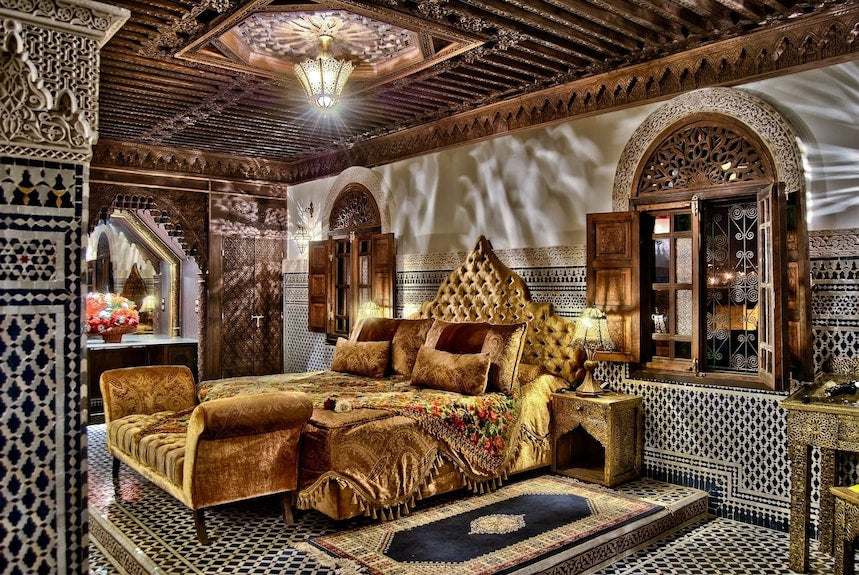 Riad Salam Fes bedroom with ornate carved wood ceiling, patterned tile walls, royal velvet upholstered bed and matching bench