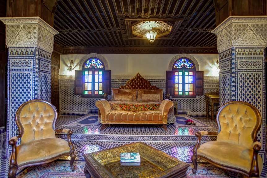 Traditional Moroccan bedroom in Riad Salam Fes with velvet tufted armchairs, patterned tile floor, carved wood ceiling, and stained glass windows