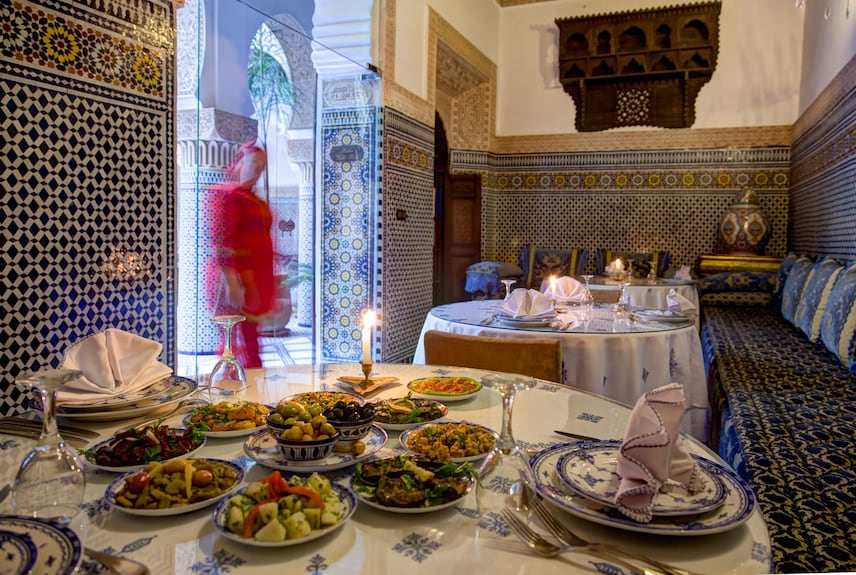 Riad Salam Fes traditional Moroccan dining room with blue and white tilework, patterned cushions, arched doorway, and a table set with colorful dishes and candles