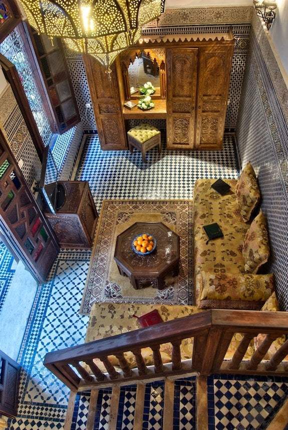 Riad Salam Fes traditional Moroccan living room with patterned tile floor, carved wooden furniture, floral sofa, and hanging lantern