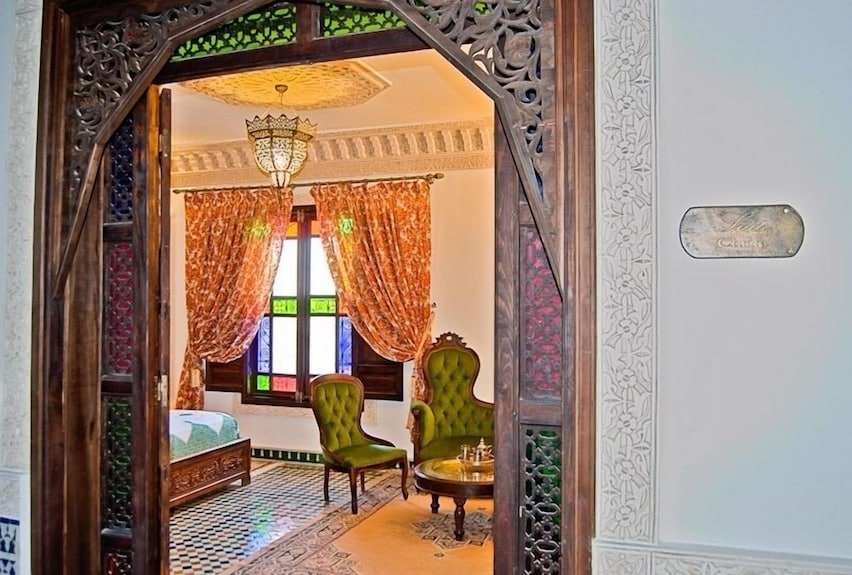 Riad Salam Fes Moroccan-style room with carved wood archway, green velvet chairs, colorful stained glass window, and patterned tile floor