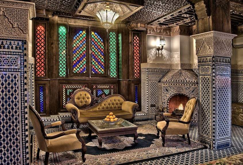 Riad Salam Fes lounge with traditional Moroccan tilework, wooden stained glass windows, ornate columns, vintage upholstered seating, and a tiled fireplace