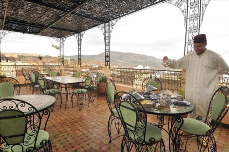 Riad Salam Fes outdoor terrace with green cushioned wrought iron chairs, tiled floor, and man serving tea under ornate canopy
