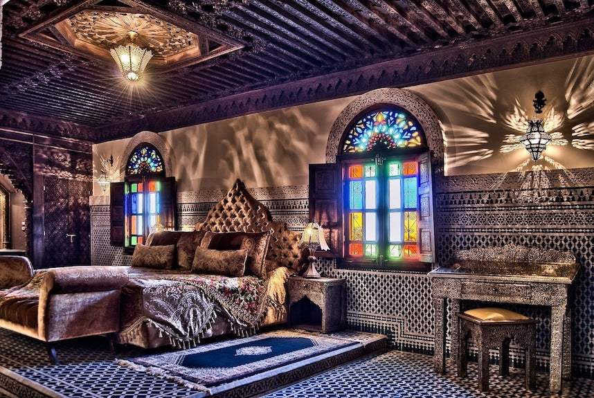 Riad Salam Fes bedroom with traditional Moroccan wooden ceiling, colorful stained glass windows, ornate patterned walls, and a plush bed with decorative pillows and textiles