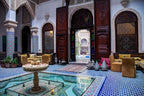 Traditional Moroccan riad interior at Riad Salam Fes featuring intricate blue and white tile flooring, wooden arches, stained glass windows, and ornate fountain in courtyard seating area