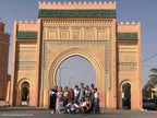Large ornate Moroccan archway in peach and green with intricate patterns, holding a group of tourists in front, representing Merzouga Sahara Desert.