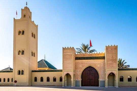 Moroccan traditional desert palace with tan walls, arched wooden doors, and a tall minaret under clear sky in the Merzouga Sahara Desert.