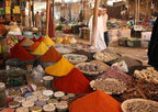 Open market stall with vibrant colorful spice piles and various herbs in baskets, bustling with people in traditional attire, reflecting the lively atmosphere of the Merzouga Sahara Desert.