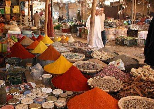 Open market stall with vibrant colorful spice piles and various herbs in baskets, bustling with people in traditional attire, reflecting the lively atmosphere of the Merzouga Sahara Desert.