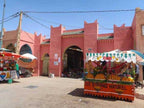 Market scene in front of pink mud-brick buildings with an orange juice cart and striped canopy, capturing a vibrant Merzouga Sahara Desert atmosphere.