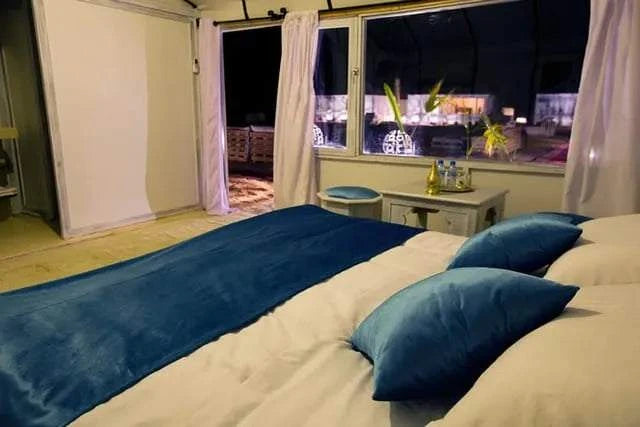 Royal Desert Camp bedroom with large bed, blue bedding and pillows, white curtains, and a nightstand with decor by a large window.
