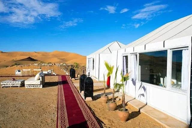 Royal Desert Camp with white canvas tents, large windows, outdoor seating, potted plants, and a red carpet walkway in the desert.