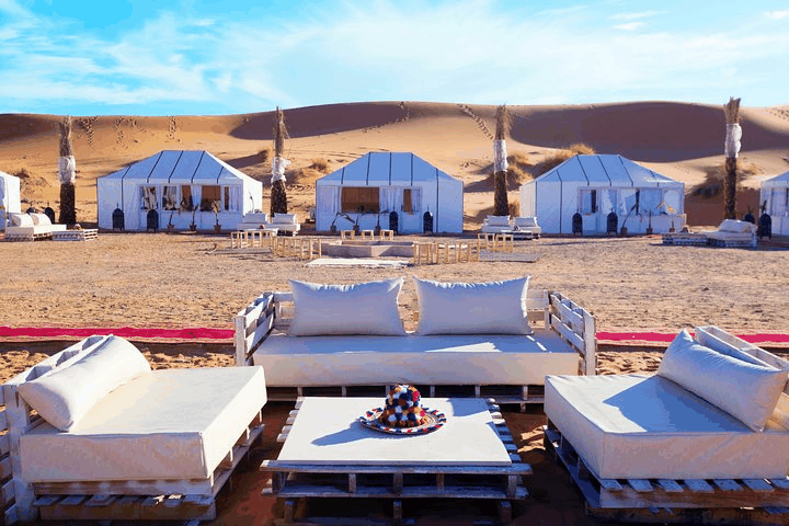 Luxury outdoor seating area with white cushions and low wooden tables in front of white canvas tents at a Royal Desert Camp in the desert.
