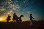 Sunset desert scene with silhouettes of camels and riders led by a guide in traditional attire at Royal Desert Camp.