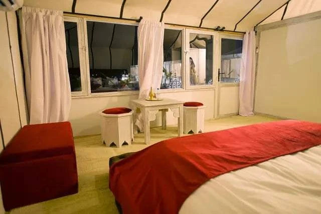 Royal Desert Camp interior with beige fabric walls, large windows, red and white bedding, and traditional Moroccan white wooden furniture.