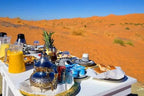 Outdoor breakfast setup with traditional Moroccan teapot, patterned cups, glasses, bread, and juice glasses on a white table in the desert, highlighting a Royal Desert Camp experience.