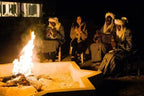 Royal Desert Camp night scene with four people in traditional clothing sitting around a fire pit, two playing musical instruments.