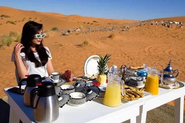 Outdoor breakfast setup at a Royal Desert Camp featuring black and white patterned ceramic dishes, fresh juice, and traditional Moroccan teapots in the Sahara sand dunes.