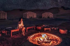 Royal Desert Camp night scene with traditional tents, starry sky, glowing campfire, and a woman sitting on wooden bench in desert sand.