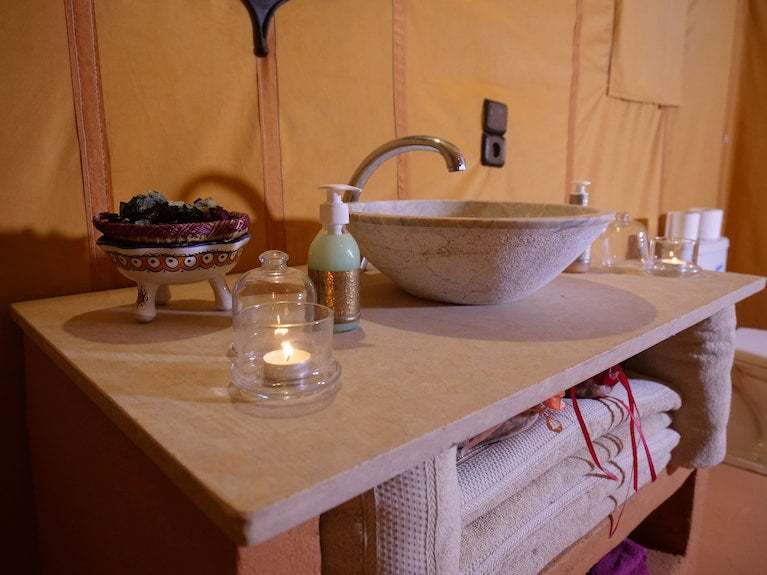 Royal Luxury Camp bathroom countertop with beige stone sink, soap dispenser, scented candle, and decorative bowl on tan surface