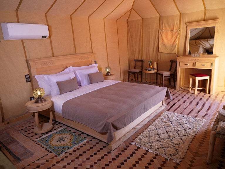 Royal Luxury Camp bedroom with beige fabric walls, large wooden bed with brown and white bedding, patterned rugs, and wooden furniture