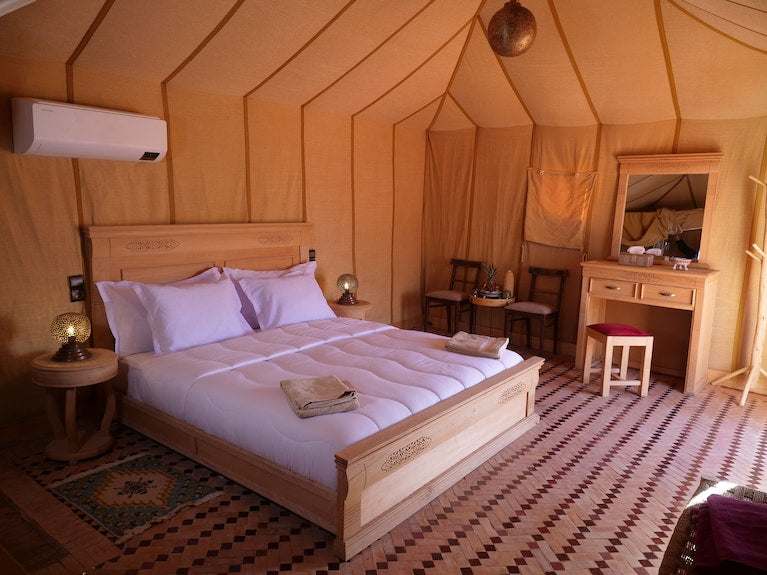 Royal Luxury Camp bedroom with wooden bed frame, white bedding, beige fabric walls, bedside tables, and vanity