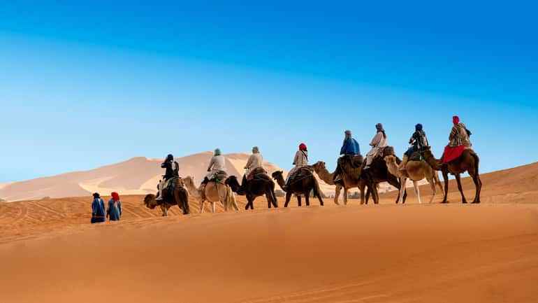 Royal Luxury Camp camel caravan with riders dressed in colorful robes trekking across golden sand dunes under clear blue sky