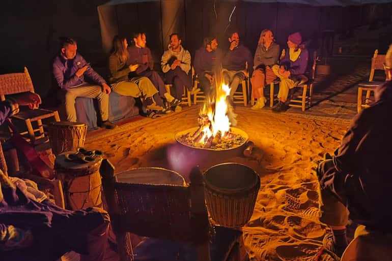 Royal Luxury Camp campfire night scene with people sitting around a glowing fire pit on desert sand seating on wooden chairs