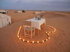 Royal Luxury Camp setup featuring a romantic dining table with white tablecloth and chairs surrounded by candles in a desert sand heart shape