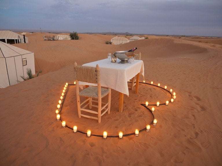 Royal Luxury Camp setup featuring a romantic dining table with white tablecloth and chairs surrounded by candles in a desert sand heart shape
