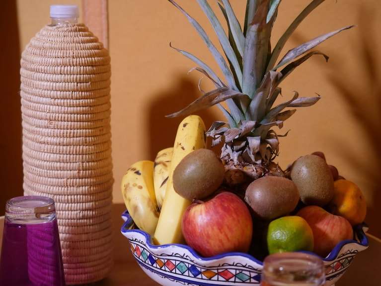 Royal Luxury Camp fruit bowl featuring pineapple, bananas, apples, kiwis, and a lime beside a woven water bottle and glass