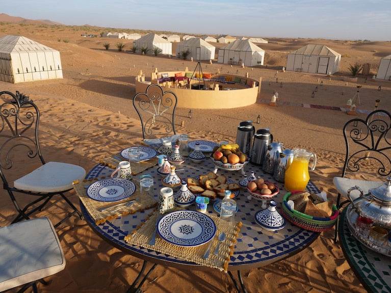 Royal Luxury Camp outdoor dining table set with blue and white patterned plates, cups, fresh fruit, bread, and juice in desert tent area