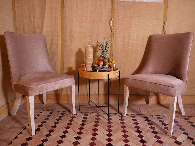Royal Luxury Camp interior featuring two light brown upholstered chairs, a round wooden side table with black metal legs, and a decorative fruit basket on a patterned floor mat