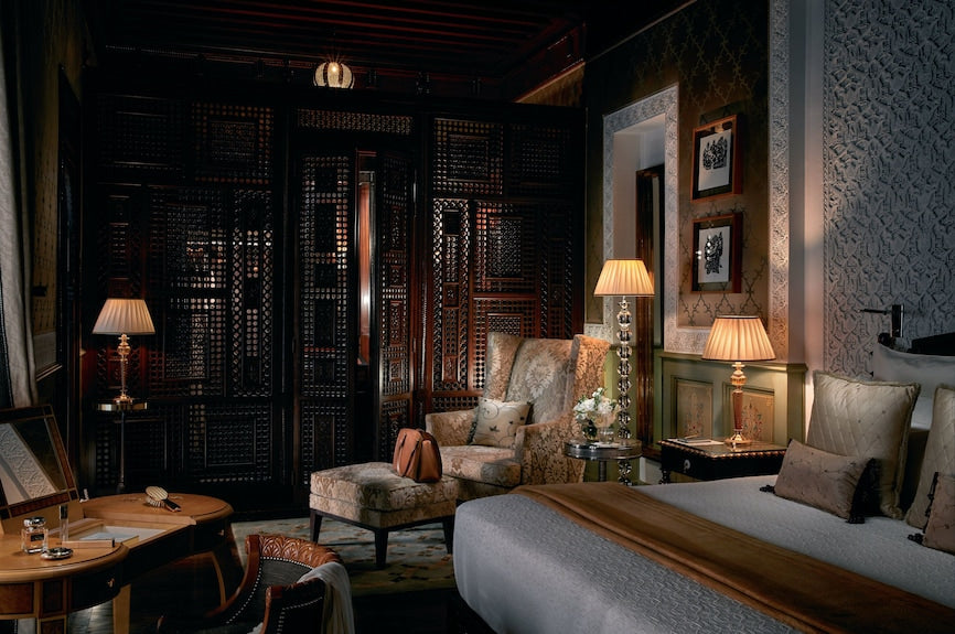 Royal Mansour Marrakech luxury bedroom with ornate dark wood screen, beige upholstered armchair, warm lighting, and patterned decor
