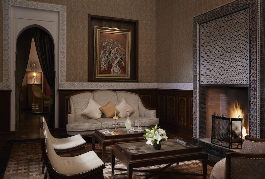 Luxury living room at Royal Mansour Marrakech featuring beige patterned sofa, wooden tables, lit fireplace with intricate tilework, and Moroccan decor