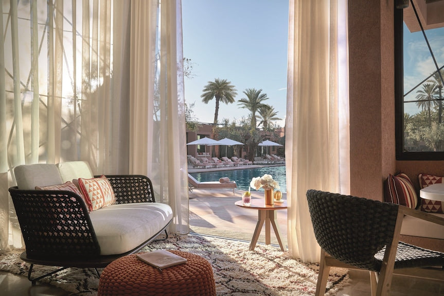 Luxury seating area with woven black frame sofa and chairs, beige cushions, and soft curtains overlooking a pool with palm trees at Royal Mansour Marrakech