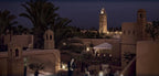 Royal Mansour Marrakech terracotta-colored buildings with traditional Moroccan architecture and palm trees at dusk with illuminated minaret in background