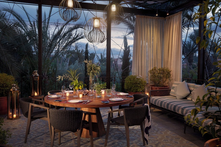 Outdoor dining area at Royal Mansour Marrakech featuring a round wooden table set with glassware and surrounded by wicker chairs and cushioned sofa under hanging lanterns