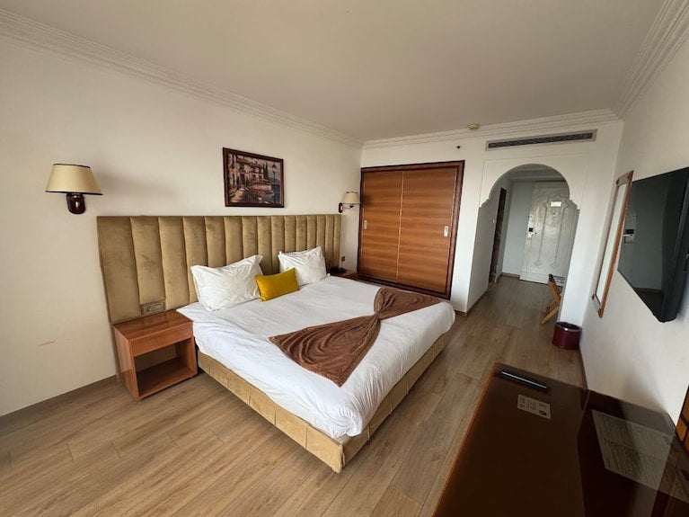 Royal Mirage Agadir hotel bedroom with white bed, beige upholstered headboard, yellow accent cushion, and wooden furnishings