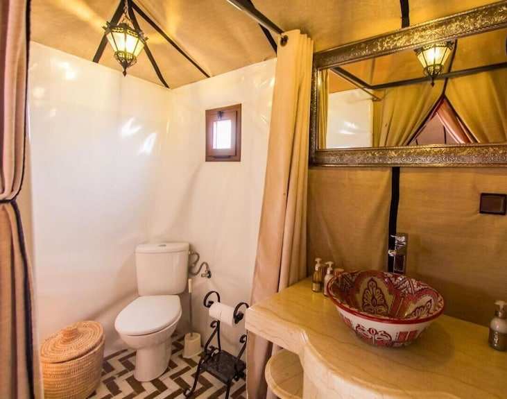 Bathroom interior at Sahara dunes Luxury Camp featuring beige fabric walls, decorative ceramic sink, toilet, and woven basket