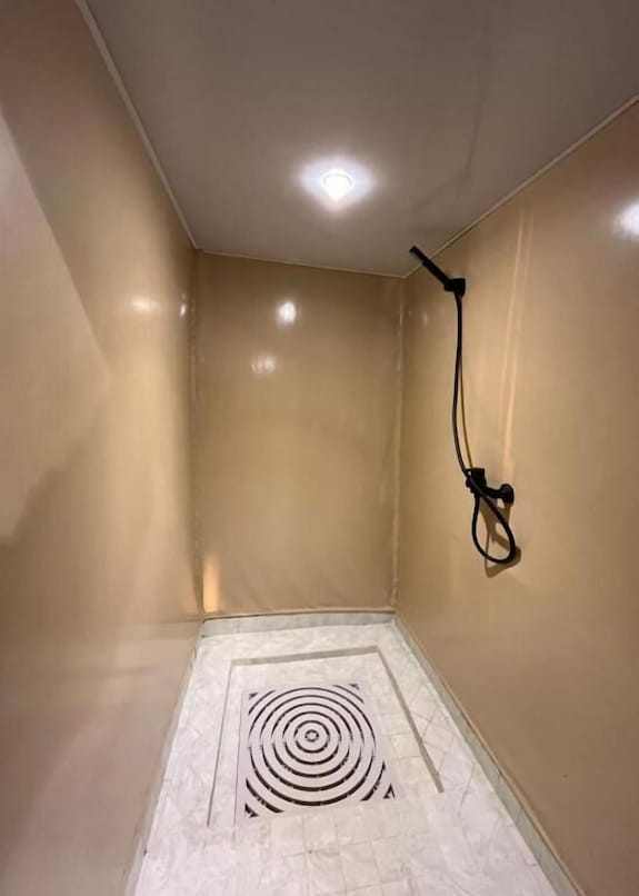 Shower area with beige walls and ceiling, white tiled floor with circular drain, and black handheld shower head at Sahara dunes Luxury Camp