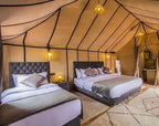 Sahara dunes Luxury Camp tent interior with beige fabric walls, two grey tufted beds with white and beige bedding, soft rugs, and warm lighting