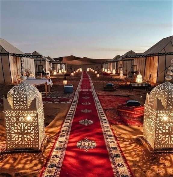 Outdoor luxury tents in beige canvas with illuminated ornate lanterns and red patterned carpets along a sandy path at Sahara dunes Luxury Camp