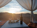 Sahara dunes Luxury Camp setup with a white cloth-covered table, wine bottle and glasses, colorful pillows, and desert sunset view