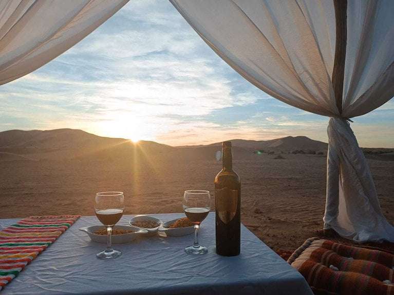 Sahara dunes Luxury Camp setup with a white cloth-covered table, wine bottle and glasses, colorful pillows, and desert sunset view