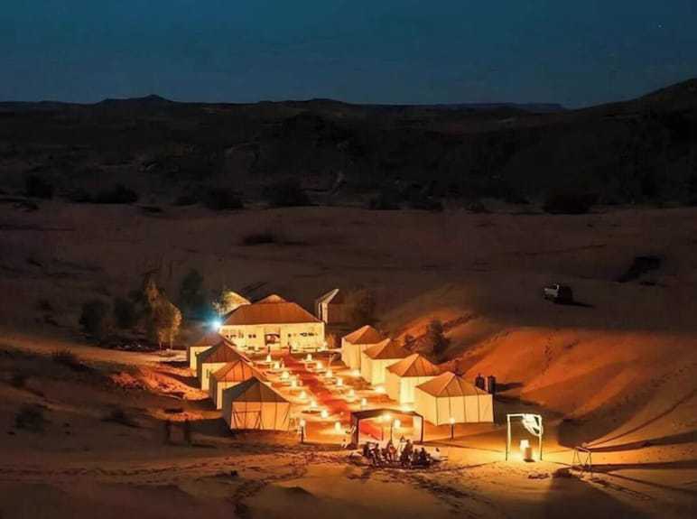 Sahara dunes Luxury Camp illuminated desert tents at night with warm lighting and a central gathering area in sandy dunes