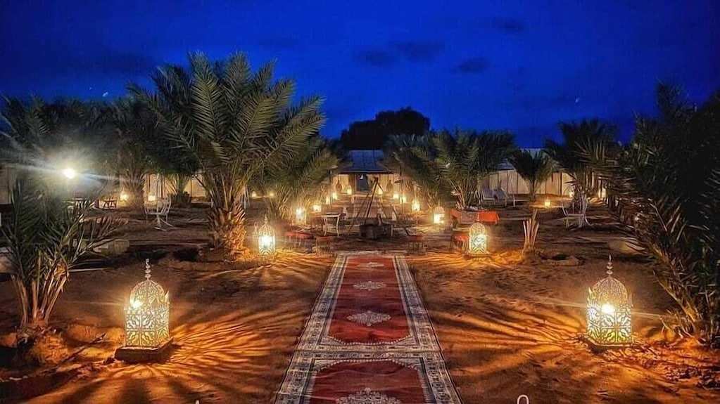 Sahara dunes Luxury Camp outdoor area at night with illuminated patterned lanterns lining a central carpet pathway surrounded by palm trees